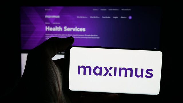 Stuttgart, Germany - 12-17-2025: Person holding mobile phone with logo of US government services company Maximus Inc. on screen in front of web page. Focus on phone display.