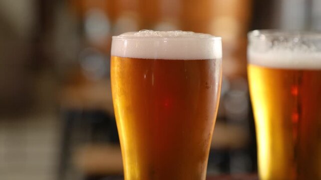 A close-up shot shows two pints of beer with foamy heads. One pint is in sharp focus while the other is blurred in the background. The setting suggests a bar or pub environment.