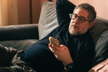 Fototapeta premium Middle aged man relaxed at home using smartphone on sofa for digital services