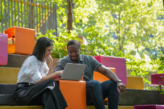 Diverse business collaboration: Black businessman and mixed-race businesswoman meeting outdoors discuss corporate document at a modern glass building green environment