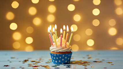 A birthday cupcake with lit candles and colorful sprinkles on a blue surface against a golden bokeh background
