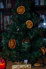 A close-up of a dried orange slice hanging as a festive ornament on a lush green Christmas tree. The slice is beautifully dehydrated, with rich amber-orange hues, translucent segments.