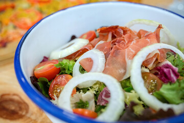 Detail of prosciutto salad mixing fresh green lettuce, cherry tomatoes, and onion rings in a white enamel bowl, on natural wood table in vegan restaurant. Organic and healthy fast food concept.