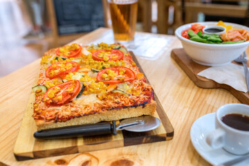 Rectangular vegetarian pizza with melted cheese, tomato, zucchini, and corn kernels, being served with a pizza cutter, a fresh poke bowl, and a coffee on a wooden table inside of italian restaurant
