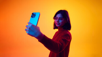 Woman taking selfie with smartphone in neon studio lighting. Concept of social media advertising, mobile camera promotion, content creation marketing, and app branding.