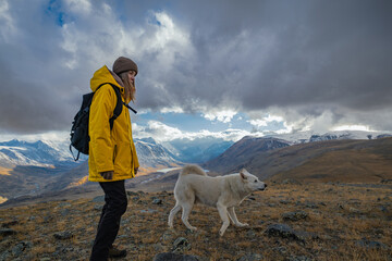 A female hiker in bright gear and her large white dog stand atop an autumn hill during golden hour. Wide side view captures them overlooking a valley with snow-capped peaks.