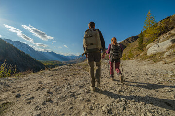 A hiking couple with backpacks and poles walks a mountain trail during golden hour, their long shadows cast on the path. Wide rear view frames them against blue skies and peaks.