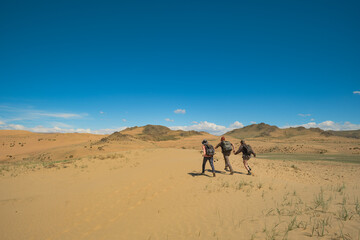 Three friends run down a sand dune in the Mongolian desert. A long-distance shot captures the joyful moment against mountains, blue sky, and patches of rare grass in the sand.