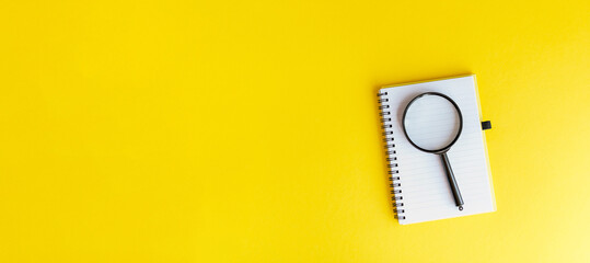 White notebook with spiral binding and magnifying glass on yellow background, minimal workspace concept.