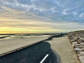 Two people walking on seaside promenade at sunset in Berck, France, along the Authie Bay, overlooking low tide sand flats and tidal channels, dramatic cloudy sky, coastal tranquility