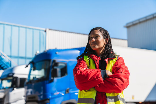 Confident woman working as a professional truck driver, standing in front of semi trailer trucks, wearing a safety vest