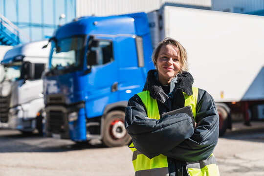 Female semi truck driver standing confident with crossed arms in front of fleet trucks at a cold storage warehouse