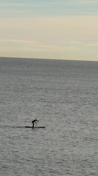 Vertical video of stand up paddle surfer moving across open sea with sailboat sailing in the distance, minimalist ocean scene focused on motion, sport, freedom and balance with calm water and copy spa
