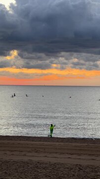 Sunset beach landscape with dramatic storm clouds over calm sea, workers cleaning sand in the foreground and paddle surfers on the horizon, environmental care concept, coastal nature scene and cinemat
