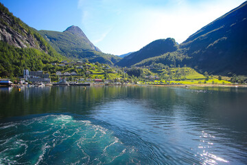 Beautiful view of Geiranger town from the water on a summer day. Reflections of the mountains in the fjord. lush green landscape with peaks towering over the village. Norway.