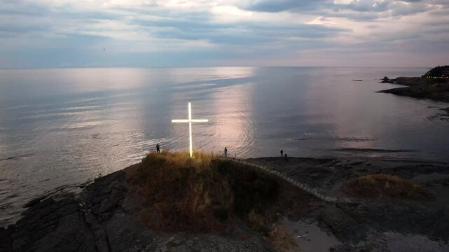Aerial view of a glowing cross on a rocky outcrop overlooking the serene sea under a cloudy sky, creating a striking contrast of light and shadow, Lozenets, Tsarevo, Bulgaria.