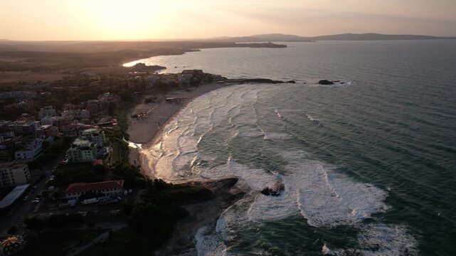 Aerial view of the coastline with the sun setting over the horizon, creating a warm glow on the buildings and the waves of Lozenets, Tsarevo, Bulgaria.