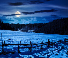 rustic wooden fence through snow covered hill at night. beautiful view in winter mountains in full moon light. alpine rural landscape near forest. finance investment or abstract concepts