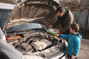 A bearded man in a windbreaker with his young son drives a car. They open the hood and pour in antifreeze, windshield washer fluid, and oil. The parent teaches his son how to operate and repair a car. © illustrissima