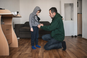 A father teaches his son how to dress and helps him zip up his jacket at home before going outside.