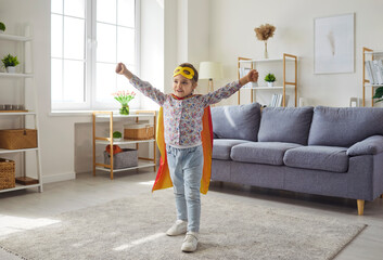 Portrait of a little cute child girl wearing super hero costume with face mask and cloak having fun standing in the living room at home. Happy childhood, home leisure and people concept. © Studio Romantic