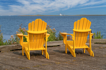 A pair of Adirondack chairs on a wooden deck overlooking the sea.