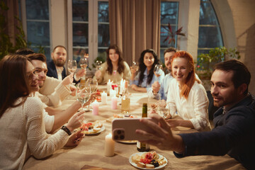 Friends raising wine glasses for group selfie at dinner. Concept of social media lifestyle content,...