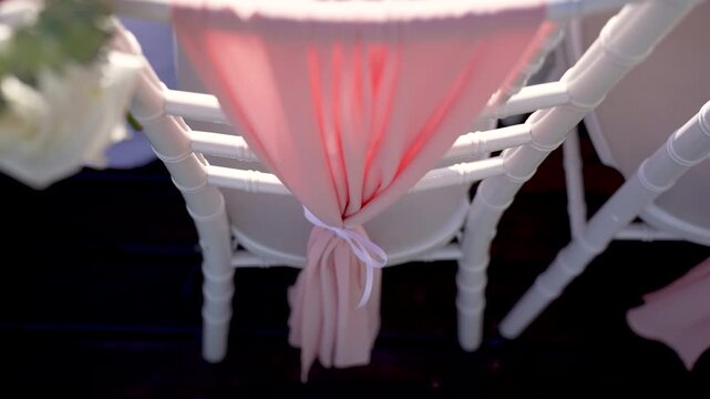 A person ties peach-colored fabric sashes around white chairs at a garden setting. Flowers are arranged nearby while the sunlight brightens the area. Guests will arrive soon.