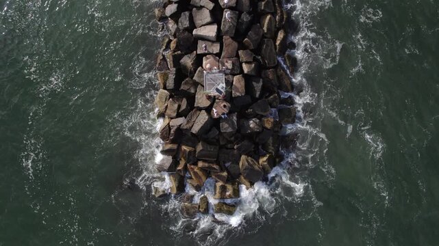 Aerial view of the dark rock breakwater meeting the dark green ocean water with white waves crashing against the rocks, Wells, Maine, United States.