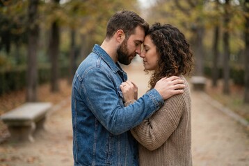 Fototapeta premium A couple stands closely together in a park during autumn, with their foreheads touching. The path is lined with trees shedding leaves, creating a serene atmosphere perfect for intimate moments