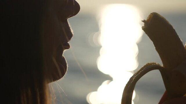 Woman eating banana fruit at sunset silhouette close up by the sea showing healthy lifestyle and diet.