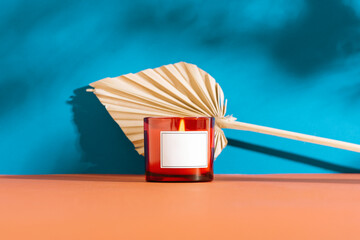 Burning red glass candle with blank label and dried palm leaf on blue and coral background in sunlight. Mockup