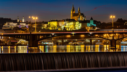 Obraz premium Bridges over the Vltava River with traffic during the ending blue hour. In the background, the illuminated Prague Castle, in the foreground, a massive river weir, underlining the whole scene