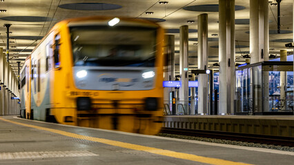 Obraz premium A small yellow regional train departs from a small modern Prague railway station. View from platform level - low perspective. The dynamics of the movement are captured by a slight blurring.