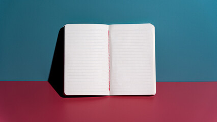 A top-view shot of an open white notebook with lined pages, placed against a vibrant dual-tone teal and pink background. Perfect for educational and creative concepts. © Andik