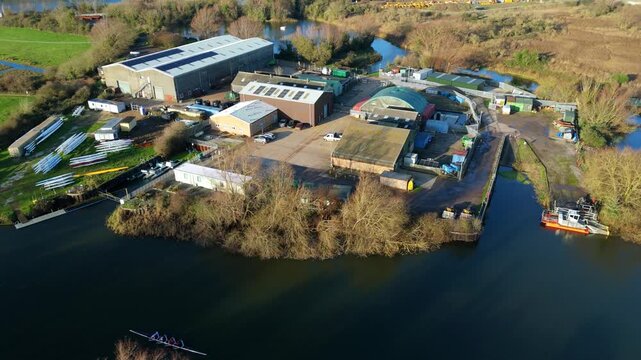 Aerial view of various buildings, boats, and bare trees along the riverbank showcasing a blend of industrial and natural landscapes, Ely, England, United Kingdom.
