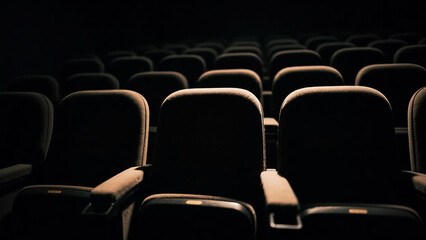 Fototapeta premium Empty rows of dark cinema seats in a movie theater, illuminated by a subtle light from above, creating a dramatic and quiet atmosphere.