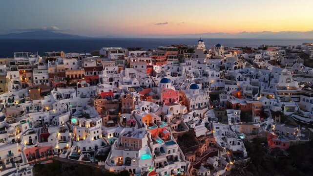 Aerial view of the dazzling white buildings of Oia cascade down the cliffs, lit by the warm glow of a setting sun, Oia, Thira, Greece.