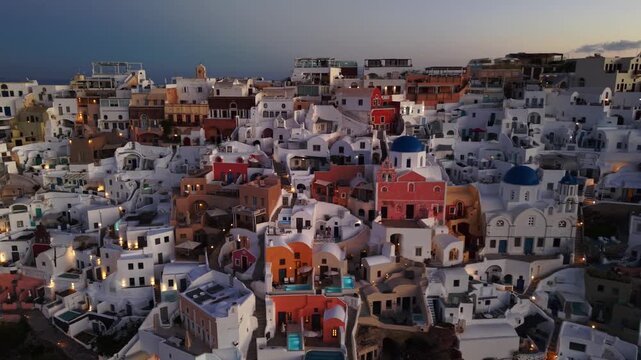 Aerial view of Oia's iconic white buildings cascading down the hillside, punctuated by vibrant blue domes against the twilight sky, Oia, Thira, Greece.