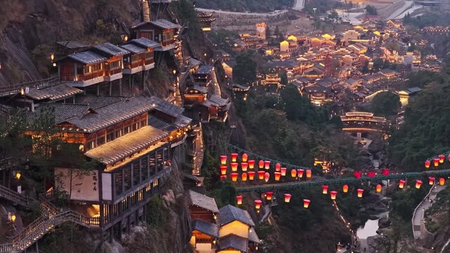Aerial view of Wangxian Valley's traditional buildings clinging to the steep cliffs, illuminated by warm lights, creating a magical ambiance, Wangxian Valley, China.