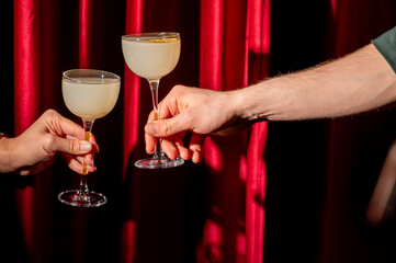 Two hands toast with elegant gin cocktails in coupe glasses against a red velvet curtain backdrop...