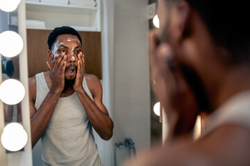 Black man looking at himself in a bathroom mirror while applying skincare around his eyes. Focused self-care routine at home, expressing concentration, calmness, and commitment to personal wellness