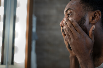 Black man looking at himself in a bathroom mirror while applying skincare around his eyes. Focused self-care routine at home, expressing concentration, calmness, and commitment to personal wellness