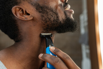 Close-up of a Black man trimming his beard with an electric shaver in a bright bathroom. Focused grooming routine at home, modern self-care habit, calm and confident mood