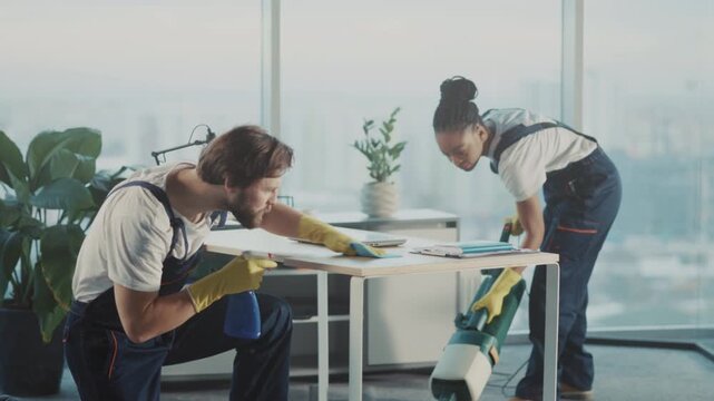 Male worker pauses cleaning process, Male cleaner momentarily rests during break as colleague vacuums by window - Powered by Adobe