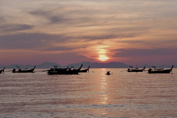 A broad line of boats floats near the horizon as golden light and soft clouds fill the sky, setting a scenic sunset scene along the water. Krabi, Thailand.