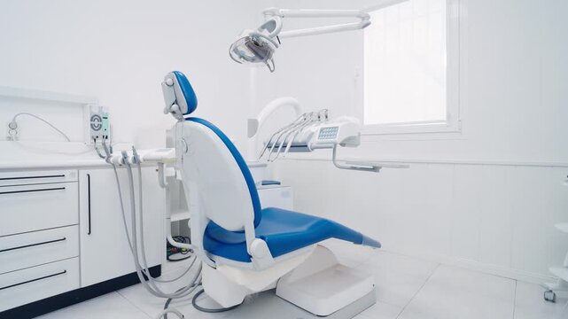 White, sterile, and modern dental clinic interior. Empty blue patient chair with professional equipment, drills, and overhead examination light