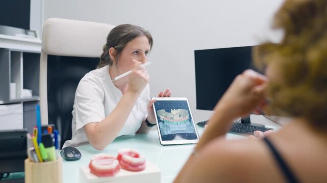 Young female dentist sitting at her desk showing a patient a digital model of a dental implant on a tablet in a modern dental clinic
