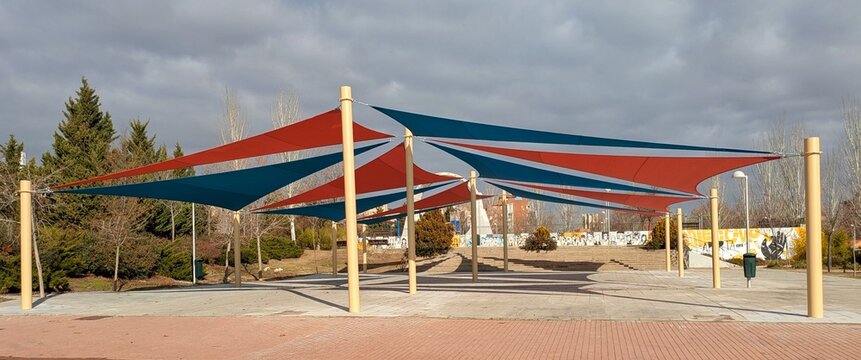 Parasoles triangulares de exterior, de color rojo y azul, sostenidos por postes en plaza p&uacute;blica 
