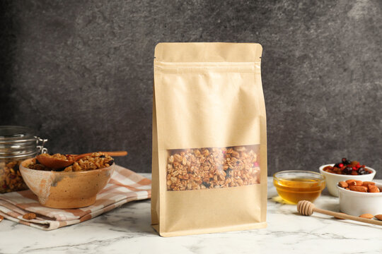 Paper bag with tasty granola on white marble table, closeup